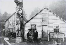 early 20th century photo of two Kwakiutl men in front of their wooden house and a totem pole